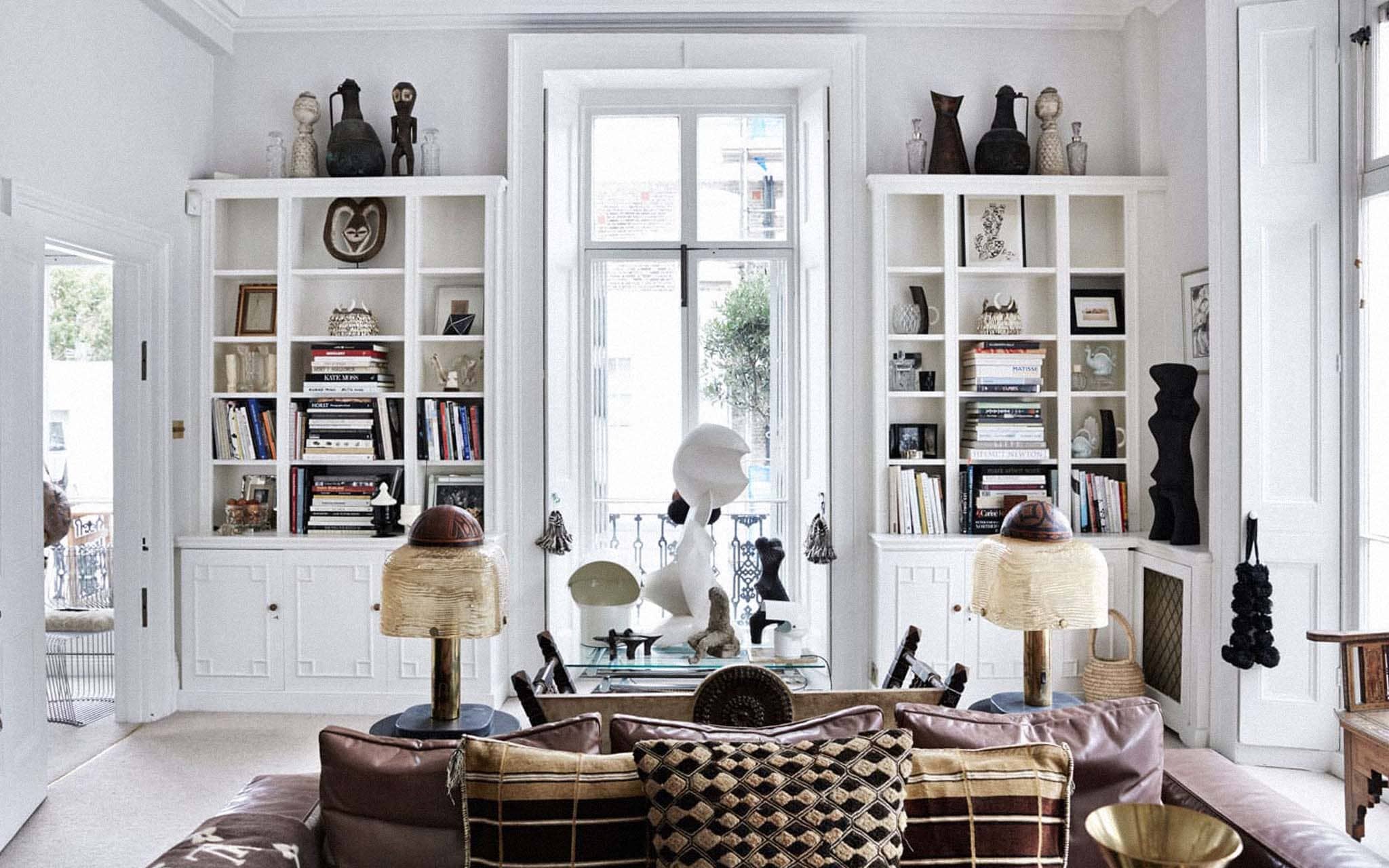 A designer lounge interior with sofa in brown leather and patterned cushions. There is a folded brown a cream patterned blanket resting on the arm. There are two matching beige and brown lamp behind the sofa with a glass table behind them with various art sculptures on it. The bookcases feature various art sculptures and vases, framed pictures and paintings, and books.